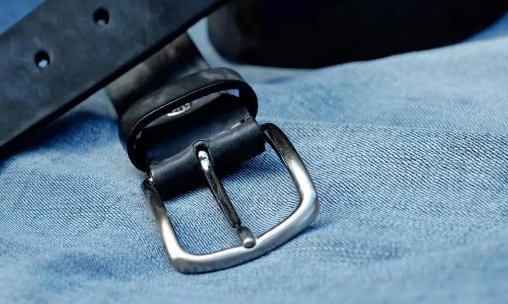 Close-up of a black leather belt with metal buckle laid on blue denim fabric.